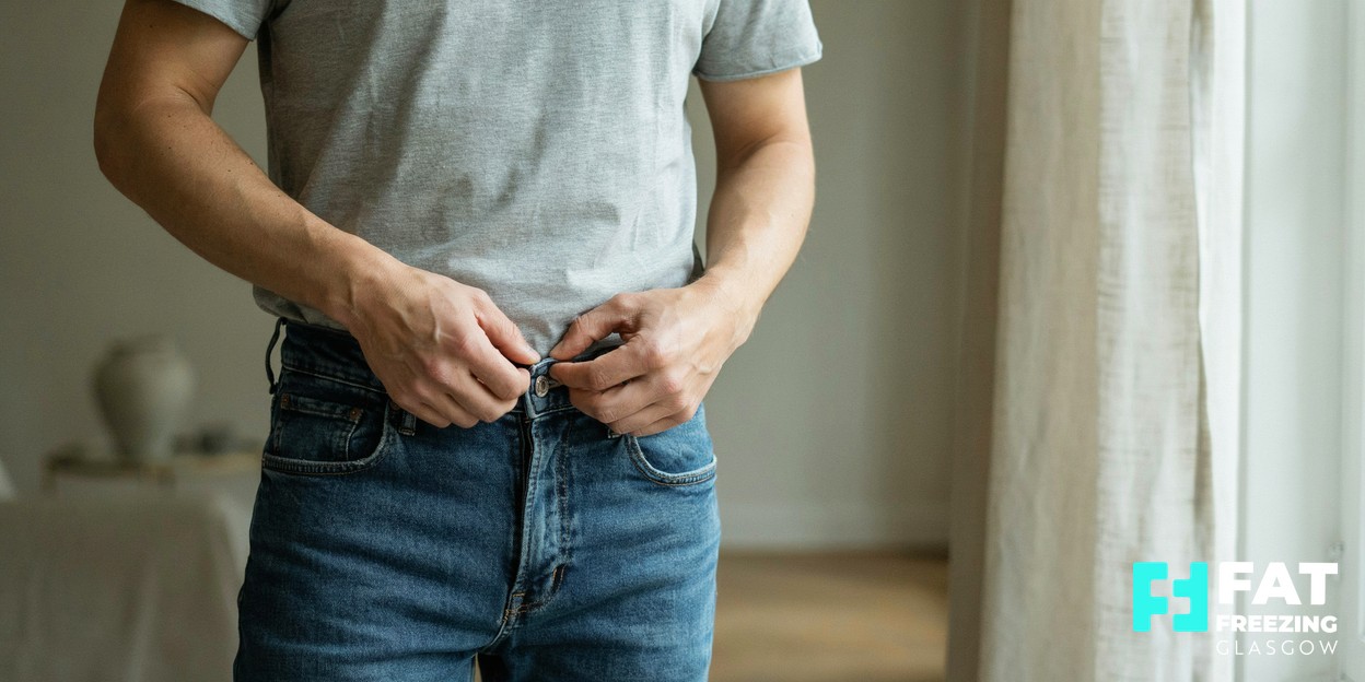 A man in a plain grey t-shirt comfortably fastening the button of well-fitting jeans in soft daylight, cropped at the waist, showing that clothing fit is the honest measure of a body-contouring result