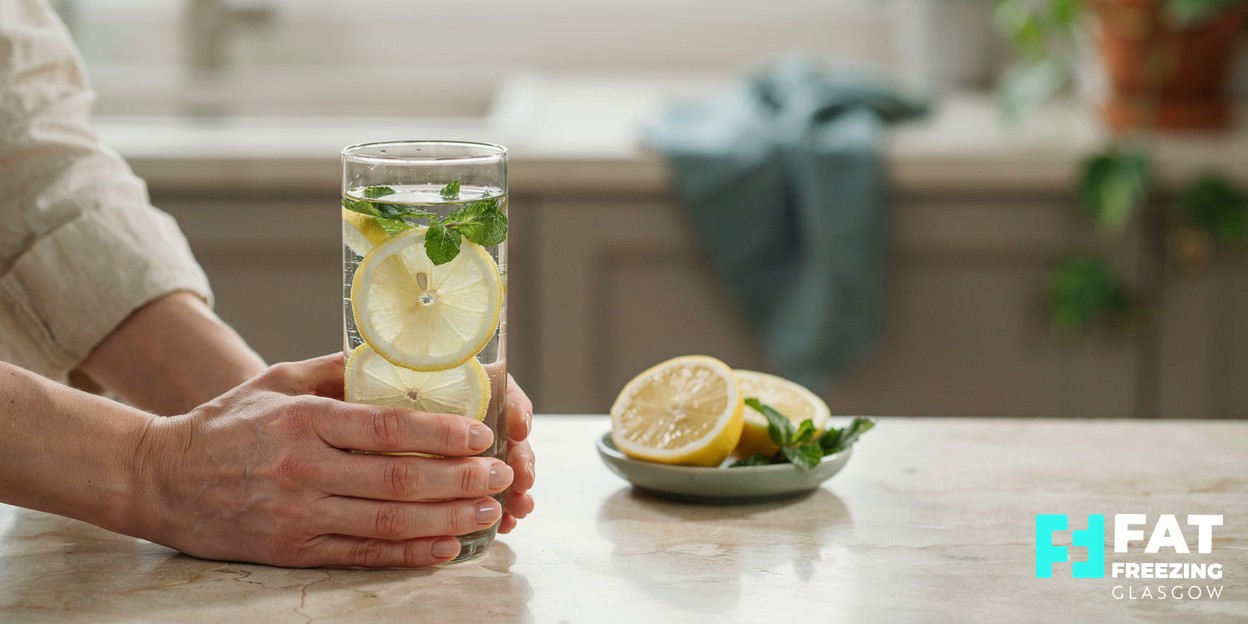 A photograph of a person’s hands cupping a clear glass of water with fresh mint and lemon on a calm kitchen counter in soft natural daylight